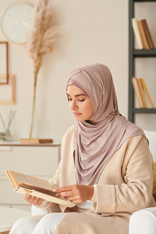 Beautiful young woman wearing stylish pale pink hijab sitting in armchair alone in living room reading novel