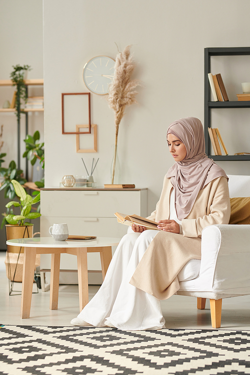 Vertical shot of modern Muslim woman wearing casual clothes spending free time in living room reading novel