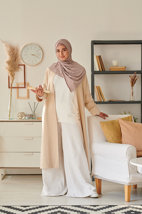 Vertical full shot portrait of attractive muslim woman wearing stylish casual outfit with hijab holding book standing in living room looking at camera