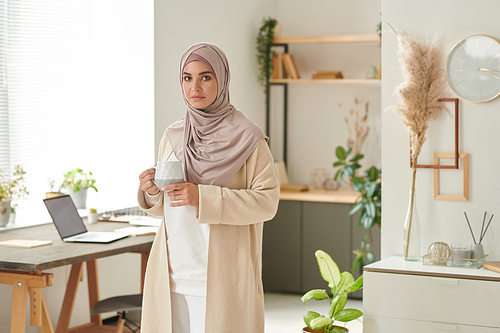 Horizontal medium ling shot of stylish Muslim woman standing alone in cozy office room holding cup of coffee, copy space