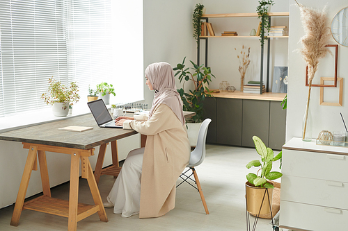 Stylish Muslim woman wearing hijab sitting alone in cozy office room working on her laptop, copy space