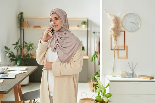 Horizontal medium long portrait of young Muslim woman wearing headscarf and casual outfit standing in modern office room talking on phone