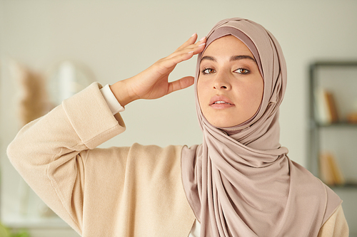 Horizontal chest up portrait of beautiful Middle Eastern woman wearing pale pink hijab touching her head with fingers looking at camera