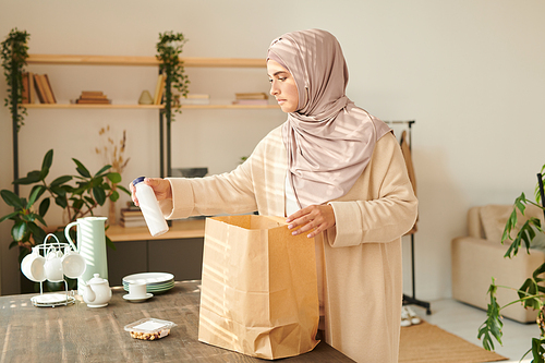 Modern Muslim woman in headscarf standing at table taking food products out of paper bag delivered to her