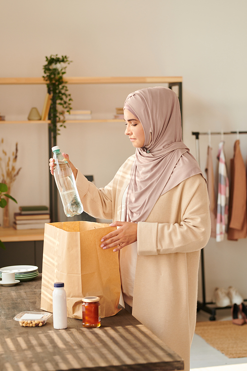 Vertical medium long shot of modern Muslim woman wearing headscarf taking out things delivered to her