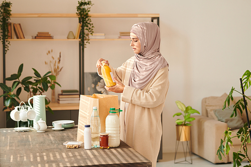 Horizontal medium long shot of young Muslim woman wearing headwrap standing at table taking out food delivered to her