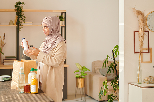 Horizontal medium long shot of cheerful young Muslim woman wearing hijab standing at table taking out food delivered to her, copy space