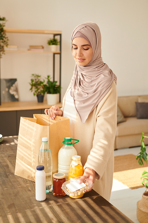 Vertical medium long shot of beautiful young Muslim woman wearing hijab standing at table taking out food delivered to her