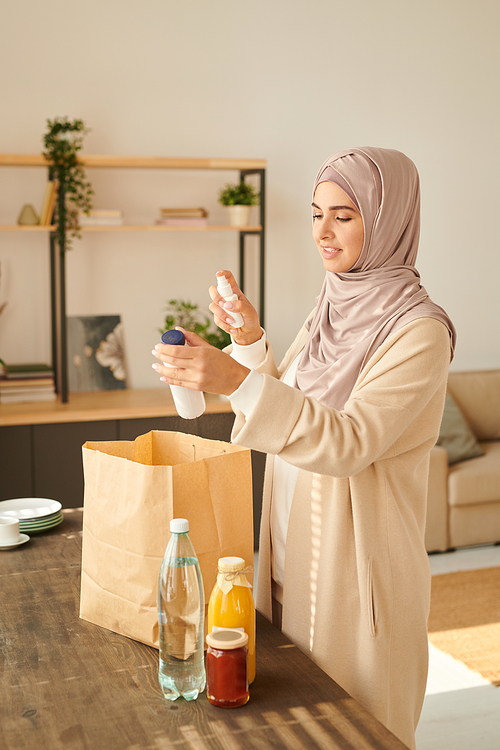 Vertical medium long portrait of modern young Muslim woman in headwrap disinfecting goods delivered to her