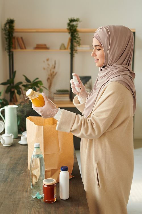 Vertical medium long side view shot of modern Muslim woman wearing protective gloves disinfecting things delivered to her during quarantine time