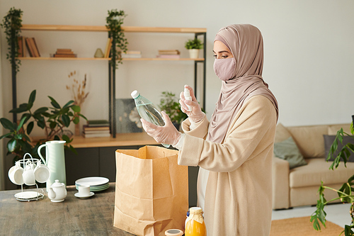 Muslim woman wearing hijab, protective mask on face and gloves disinfecting goods at home after going shopping