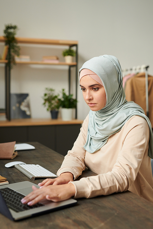 Vertical portrait of beautiful young woman wearing light blue hijab sitting at table working on laptop at home