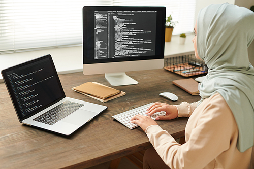 Unrecognizable young woman wearing light blue headscarf working on computer code using desktop PC