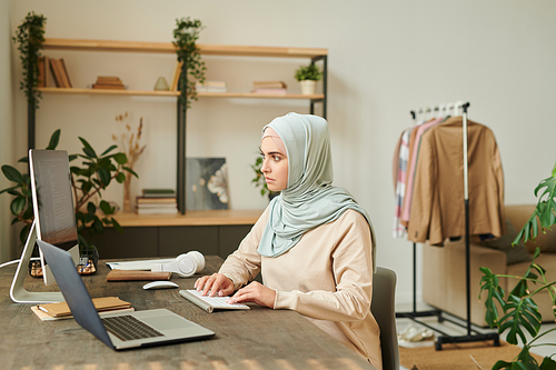 Serious young Muslim professional programmer wearing light blue hijab sitting at desk at home working on software