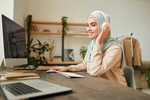 Portrait of professional software developer wearing hijab and headphones sitting at desk having online meeting while working on computer