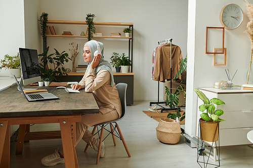 Beautiful young Muslim woman wearing hijab and headphones working remotely at home, copy space