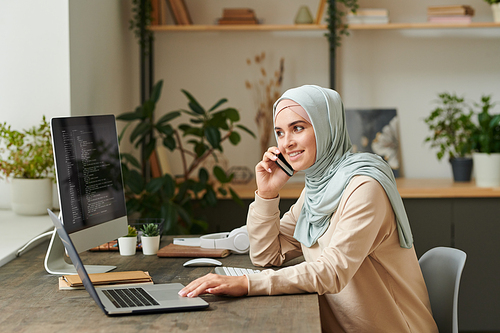 Cheerful young woman wearing light blue hijab sitting at table talking on phone while working on computer