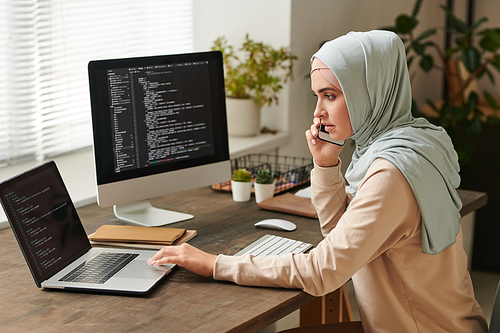 Young woman wearing light blue hijab sitting at table discussing important issues on phone while working on software