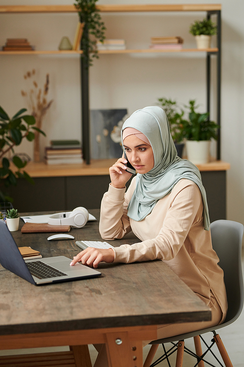 Serious young woman wearing light blue hijab sitting at table discussing important issues on phone while working on her laptop