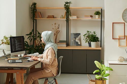 Young Middle Eastern professional programmer wearing light blue hijab sitting at desk at home working on new computer software, copy space