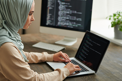 Professional programmer wearing hijab sitting at office table typing computer code on laptop, copy space