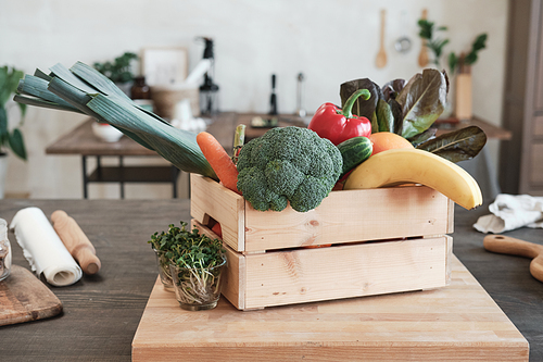 Wooden box full of various vegetables and fruits placed on board with growing plants into glasses in modern kitchen