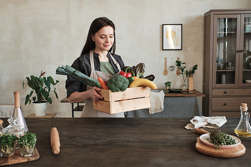 Content beautiful young woman in apron carrying wooden box of vegetables and fruits to table with rolling pin and boards
