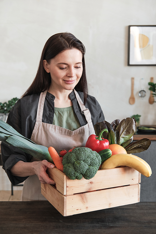 Smiling attractive young woman in apron coming with box of fresh vegetables to table while preparing ingredients for cooking