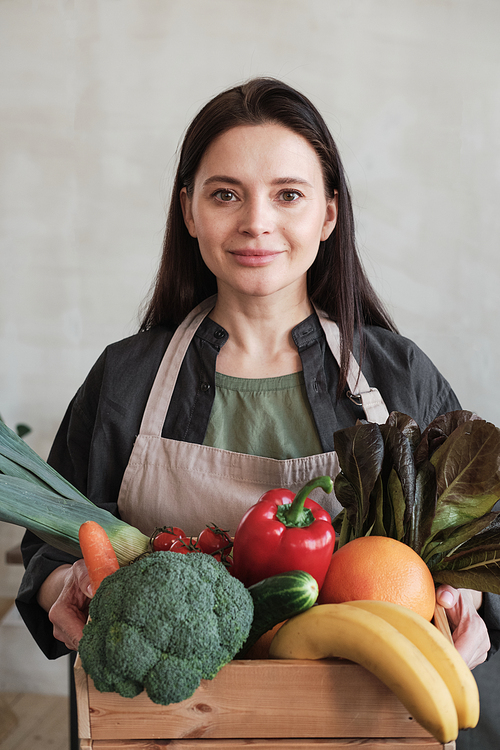 Portrait of content brunette young housewife in apron standing with box full of ripe vegetables and fruits