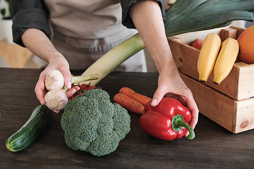 Close-up of unrecognizable woman in apron putting vegetables on table while unpacking wooden box