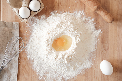 Directly above view of cracked egg into flour on wooden board with rolling pin, whisk and eggs