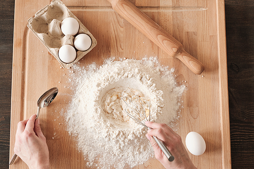 Above view of unrecognizable woman mixing flour and egg with whisk on wooden board with rolling pin