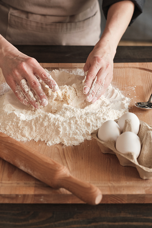 Close-up of unrecognizable woman in apron standing at kitchen counter and mixing egg with flour while making dough on wooden board
