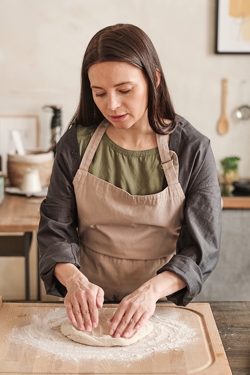 Serious young woman in apron standing at table with wooden board and kneading dough while cooking buns in kitchen