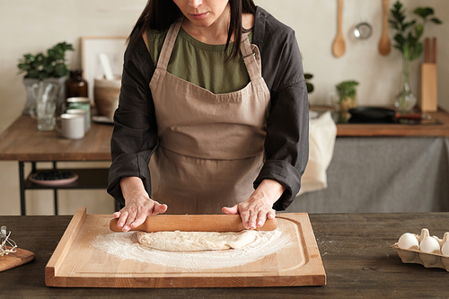 Busy woman in apron standing at table and rolling dough with pin on wooden board in modern kitchen