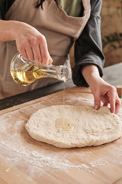 Close-up of female hand pouring oil from jug onto dough while making homemade pizza
