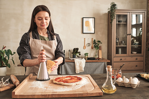 Content brunette young woman in apron standing at counter with ingredients and using grater for cheese while cooking homemade pizza