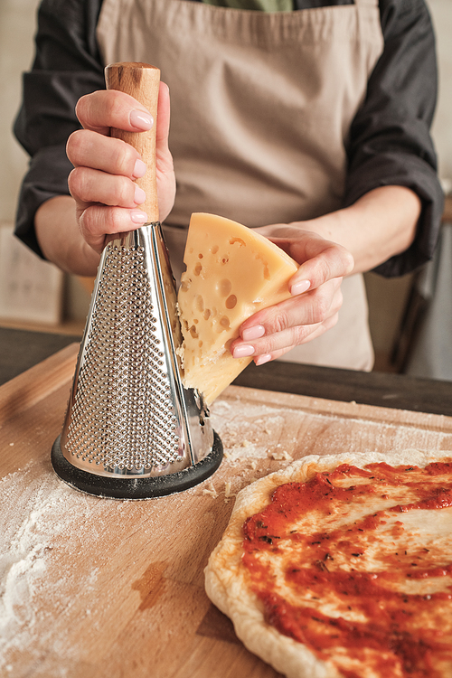 Close-up of unrecognizable woman in apron using cheese grater on wooden board with pizza dough greased with tomato sauce