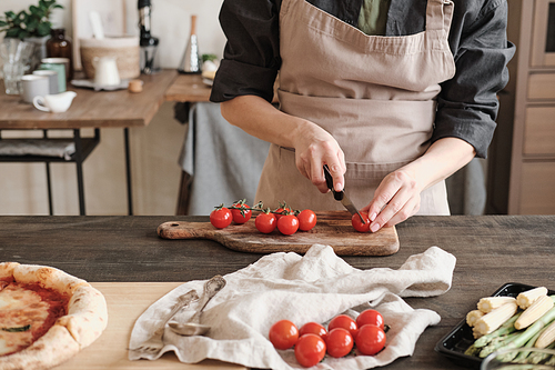 Close-up of unrecognizable woman in apron standing at counter with vegetables and cutting tomatoes on wooden board