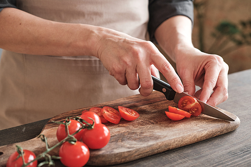 Close-up of female hands slicing cherry tomato with knife on wooden board, cooking vegetable salad