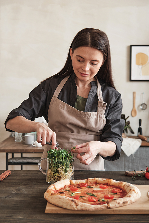 Content brunette young woman in apron standing at counter and cutting pea leaves for homemade Italian pizza