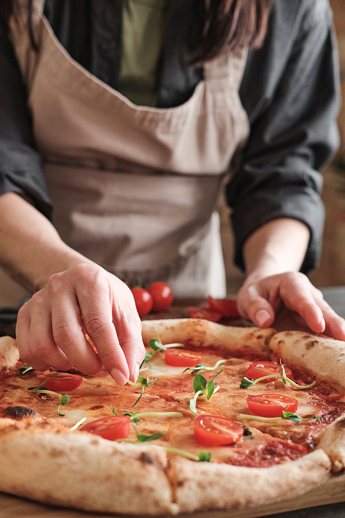 Close-up of unrecognizable woman in apron putting pea leaves on pizza with tomatoes while preparing it for family