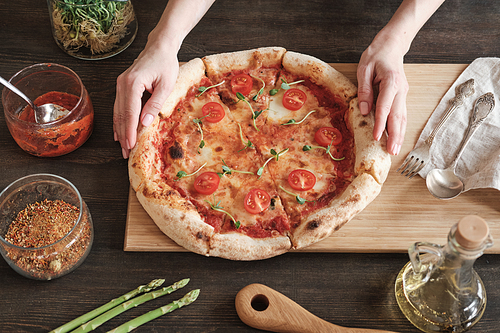 Directly above view of female hands adjusting delicious homemade pizza on wooden board surrounded with jars of spice and sauce