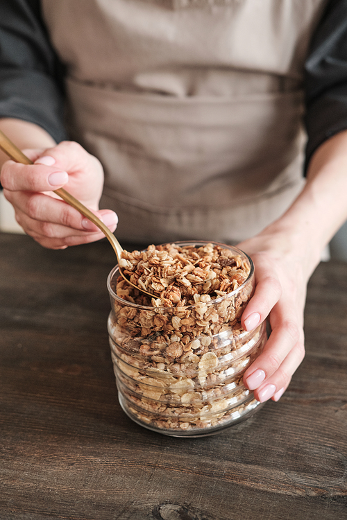 Close-up of unrecognizable woman in apron standing at wooden table and preparing granola with muesli