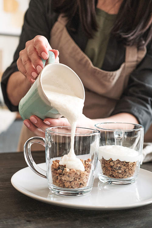 Close-up of unrecognizable woman in apron pouring yogurt into mug with muesli while making healthy dessert for breakfast