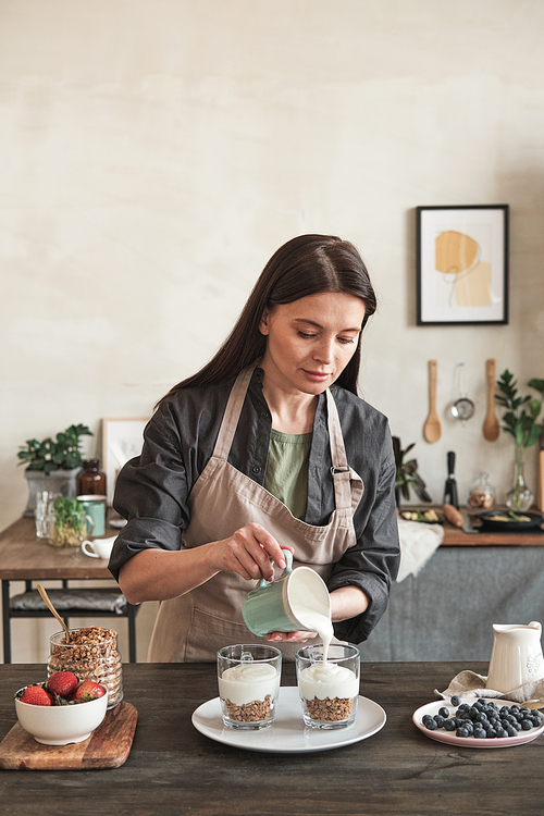 Serious brunette woman in apron leading healthy lifestyle standing at dining table and making granola with yogurt in kitchen