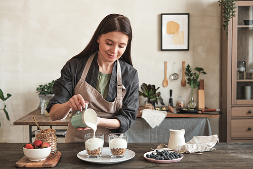 Content attractive young woman in apron standing at kitchen counter and pouring yogurt into mugs while cooking granola for healthy ration