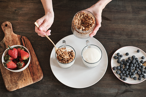 Above view of female hands holding jar and adding muesli into yogurt while making healthy breakfast