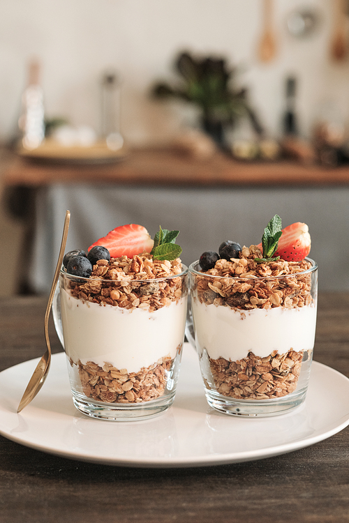 Close-up of two glassy mugs of granola decorated with berries placed on plate with golden spoon in domestic kitchen