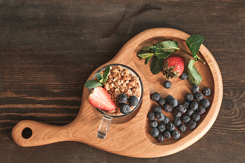 Directly above view of wooden board with granola in mug, berries and leaves on dark brown table
